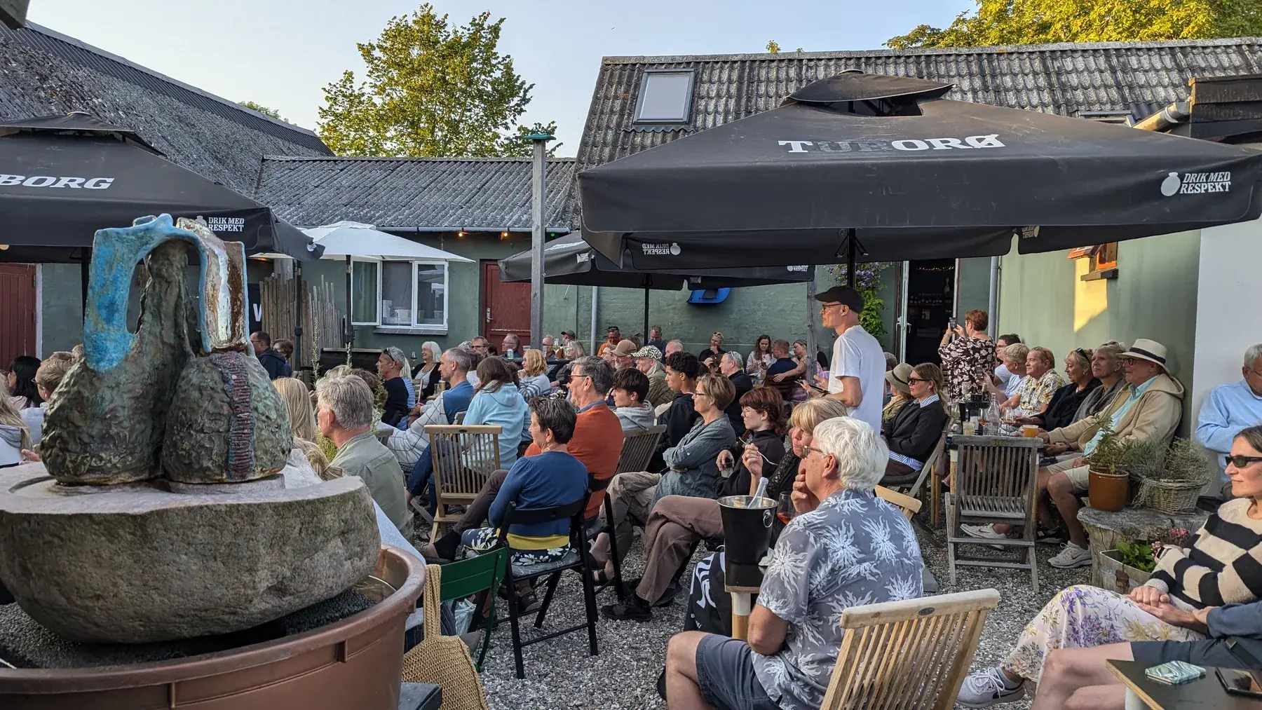 Publikum sidder under parasollerne i gårdhaven